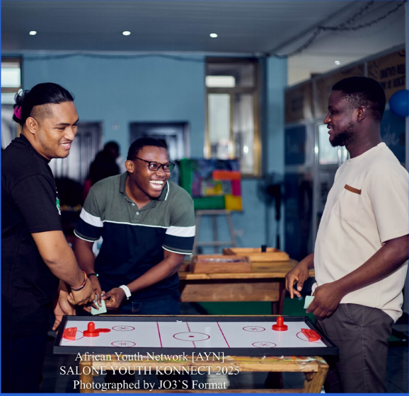 Three young men laughing over a game of air hockey at Salone Youth Konnect 2025